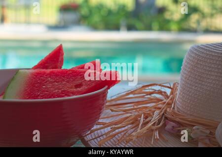 Eine Schüssel mit Wassermelone und einen Hut gegen die Sonne auf den Tisch und Schwimmbad im Hintergrund. Stockfoto