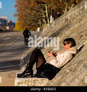 Eine Frau genießt den Winter Sonne, während auf Ihrem Telefon von den Ufern der Rhone in Lyon, Frankreich Stockfoto