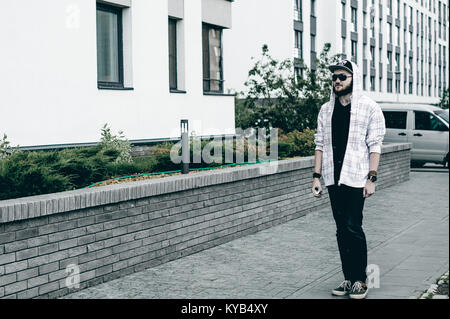 Junger Mann mit Bart in hell karierten jacke mit Kapuze in einem Hut und Sonnenbrille frei herumlaufen Stadt raucht eine e-Zigarette Stockfoto