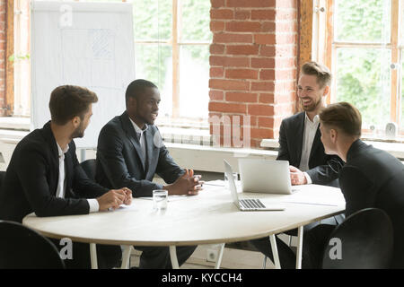 Vier fröhlichen diverse Geschäftsleute sprechen lachend an Tisch und lächelt zufrieden multirassischen Executive Group oder Partner Team diskutieren neue ide Stockfoto