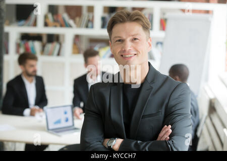 Lächelnd zuversichtlich Executive in der Klage stehen mit den Armen an Kamera mit Geschäftspartnern Team im Hintergrund gekreuzt, happy Company ceo Co-gefunden Stockfoto