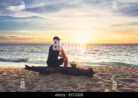 Nachdenklicher junger Mann mit Kokosnuss Drink relaxen am Strand bei Sonnenuntergang. Phuket, Thailand. Stockfoto