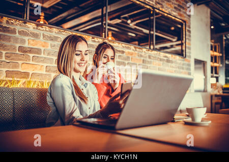 Zwei lächelnde Mädchen mit Laptop und trinken Kaffee, während in einem modernen Cafe innen sitzen. Freundschaft und Technologie. Selektiver Fokus und geringer Tiefe Stockfoto