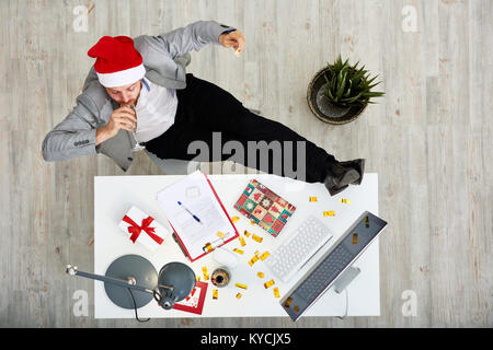 Direkt oberhalb der Blick der jungen weißen Arbeiter in Santas Hut feiern das neue Jahr bei der Arbeit: Er mit Beinen auf dem Schreibtisch und trinken Champagner aus dem Sitzen Stockfoto