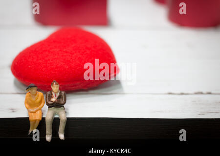 Miniatur, ein paar liebe Menschen sitzen auf einem hölzernen Tisch mit roten Herzen Hintergrund. Kopieren Sie Raum mittels wie Valentinstag Konzept, Liebe Paar Konzept. Stockfoto