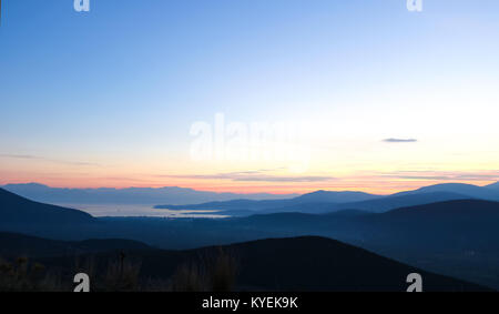 Sonnenuntergang über Blau 2-in-1-Berge und das Meer weit unten mit ein paar Lichter von Häusern und Wind turbanes auf einem fernen Hügel Stockfoto