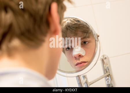 Weiße Teenager Jungen Blick auf sein Spiegelbild in einem kleinen Badezimmer Spiegel an seine Haut mit einem Streifen auf seiner Nase loswerden von Mitessern und Pickeln zu erhalten Stockfoto