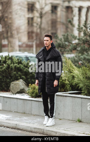 Stilvolle Mann wolk auf der Straße. Winter kalt Outfit. Große Jacke mit weißen Turnschuhen. Blured Hintergrund Straße. Profecional Modell Foto. Stockfoto