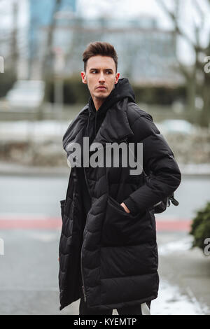 Stilvolle Mann wolk auf der Straße. Winter kalt Outfit. Große Jacke mit weißen Turnschuhen. Blured Hintergrund Straße. Profecional Modell Foto. Stockfoto