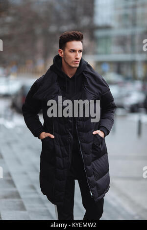 Stilvolle Mann wolk auf der Straße. Winter kalt Outfit. Große Jacke mit weißen Turnschuhen. Blured Hintergrund Straße. Profecional Modell Foto. Stockfoto