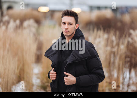 Stilvolle Mann wolk auf der Straße. Winter kalt Outfit. Große Jacke mit weißen Turnschuhen. Blured Hintergrund Straße. Profecional Modell Foto. Stockfoto