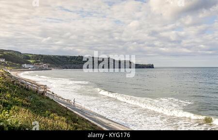 Panorama der Sandsend in der Nähe von Whitby. Wellen im Wäschetrockner in der Strand mit einer Landspitze im Hintergrund. Ein bewölkter Himmel ist Overhead. Stockfoto