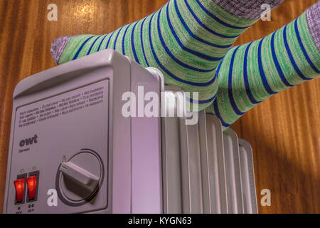 Füße von Person in ein paar grüne und blaue hooped/gestreiften Söckchen, ruht auf einem heißem Öl Heizung / Kühler, mit Power Schalter leuchten. UK. Stockfoto