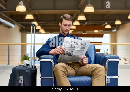 Portrait von gutaussehenden jungen Mann mit Koffer lesen Zeitung in Leder Sessel sitzt während der Wartezeit für den Check-in in Modem Hotel Stockfoto