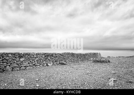 Alte Mauer aus Stein, die auf einem felsigen Strand verläuft senkrecht zur Kante des Meeres. Moody geheimnisvolle Szene. Schwere Wolken. Schwarz und Weiß. Faro, Schweden Stockfoto