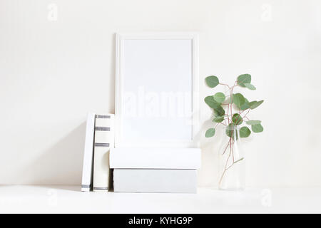 Weiße leere Holzrahmen mockup mit einem grünen Eukalyptus Niederlassungen in Glasflasche und Stapel Bücher auf dem Tisch liegen. Poster Produkt Design. Styled lieferbar feminine Fotografie. Home Decor. Stockfoto