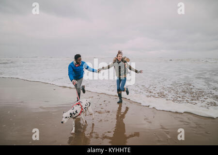 Ein junges Paar laufen weg von den Wellen, während man ihren Hund auf einem winter Strand. Stockfoto