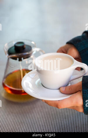 Kaukasische frau Hände halten Keramik Tasse mit schwarzem Tee und Milch, Teekanne aus Glas im Hintergrund gegenüber metallischen Tabelle Stockfoto