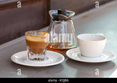 Verbrauchte cortado Kaffee in ein Glas mit Keramik weiße Tasse und Kännchen auf metallischen Tabelle und eine rostige Hintergrund Stockfoto