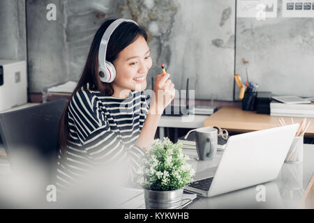 Jungen asiatischen casual Geschäftsfrau Arm auf dem Schreibtisch rest Pose mit Laptop und das Hören von Musik über Kopfhörer und über die Arbeit denken, arbeiten zu Hause mit Stockfoto