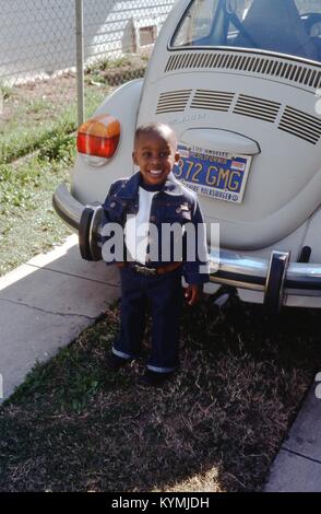 Porträt eines lächelnden African American Boy, vor einem weißen 1972 VW Volkswagen Käfer in einer Einfahrt in Los Angeles, Kalifornien, 1977 geparkt posieren. Der kleine Junge trägt eine passende Dark Wash denim Jacke und Handschellen Blue Jeans mit einem Ledergürtel und silbernen Schnalle. Der Wagen gelb-auf-blau Kalifornien Nummernschild hat eine 6-stellige Variable lesen 372 GMG, mit einem Juli 1977 Registrierung Aufkleber. Der Kennzeichenrahmen wirbt die Wilshire Volkswagen Dealership in Los Angeles. () Stockfoto