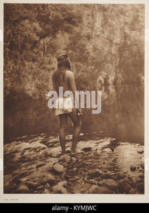 Eine historische Fotografie eines Apachen-Indianers, aufgenommen von Edward Curtis Anfang des 20. Jahrhunderts. Das Foto zeigt das Motiv in traditioneller Apachen-Kleidung, einschließlich eines Lendendeckels, das nahe einem Fluss steht und von einer natürlichen Landschaft umgeben ist. Stockfoto