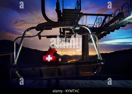 Rettungsschwimmer im roten Mantel mit medizinischen Kreuz Standortwahl auf dem Stuhl in der Seilbahn entfernt, mit Blick nacht City Light im Mountain Ski Resort Stockfoto