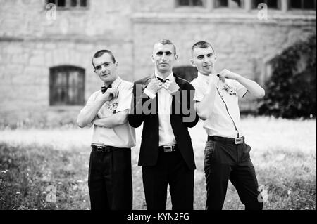 Bräutigam posiert mit zwei Trauzeugen auf einem sonnigen Hochzeitstag neben dem Schloss. Schwarz-weiß Foto. Stockfoto