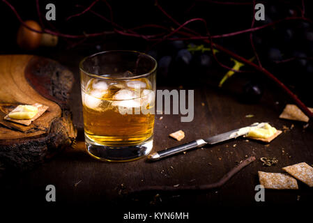 Glas Whisky mit Eiswürfel auf hölzernen Tisch serviert. Vintage Arbeitsplatte mit Trauben, Kekse und ein Glas Schnaps. Stockfoto