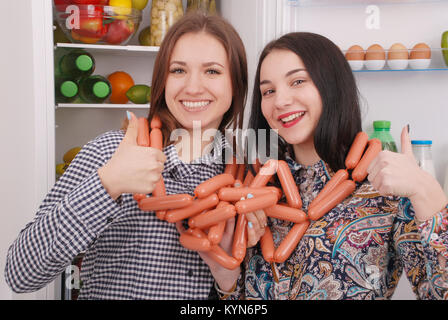 Zwei junge Mädchen hält Würstchen auf dem Kühlschrank Hintergrund. Zwei schöne junge Mädchen in der Nähe der Kühlschrank. Stockfoto