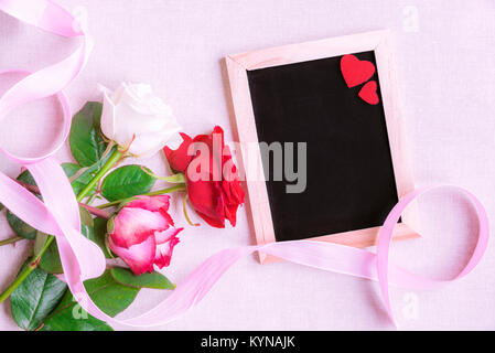 Schönen Blumenstrauß aus den vielfarbigen Rosen, mit einer rosa Schleife gebunden, in der Nähe eine leere Schiefertafel mit zwei roten Herzen dekoriert, auf einem rosa Hintergrund. Stockfoto