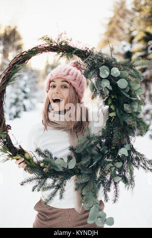 Blonde junge Dame in rosa Strickmütze halten Weihnachten Kranz mit verschneiten Winterwald auf Hintergrund Stockfoto