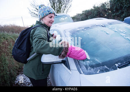 Weiße Frau mittleren Alters in einem bobble Hut mit einem rosa Handschuh Eiskratzer löscht das Eis weg Ihren silbernen Auto bevor Sie geht an einem verschneiten Morgen zur Arbeit. Stockfoto