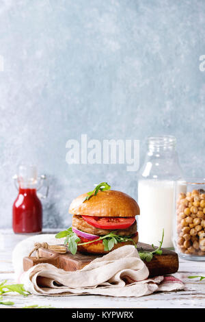 Hausgemachten Burger im klassischen Brötchen mit Tomaten, Rucola, Fleisch, Käse, Zwiebel auf Holzbrett mit einer Flasche Ketchup, Milch serviert, geröstete Kichererbsen ove Stockfoto