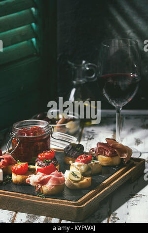 Tapas oder Bruschetta Vielfalt. Brot mit Schinken Rohschinken, sonnengetrocknete Tomaten, Olivenöl, Oliven, Paprika auf schiefer Holz mit Glas Rotwein über Stockfoto
