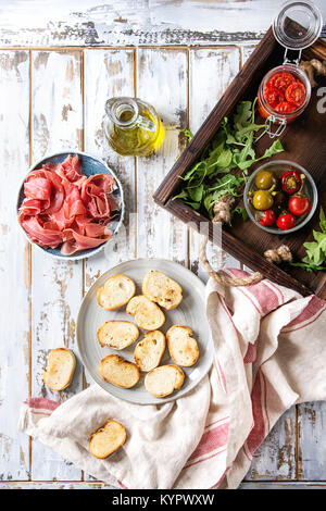 Zutaten für die Herstellung von Tapas oder Bruschetta. Knuspriges Brot, Schinken, Prosciutto, sonnengetrocknete Tomaten, Olivenöl, Oliven, Paprika, Grüns auf Platten über Weiß p Stockfoto