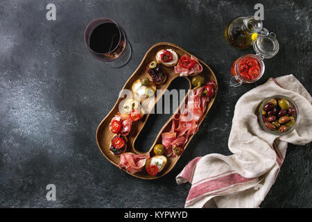 Tapas oder Bruschetta Vielfalt. Brot mit Schinken Rohschinken, sonnengetrocknete Tomaten, Olivenöl, Oliven, Pfeffer auf dekorative Holz Platte mit Glas Rotwein Stockfoto