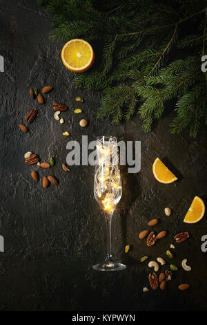 Weihnachtsbeleuchtung Girlande in Champagner Glas mit Muttern, Orange, Tanne Niederlassungen über dunkle Textur Hintergrund. Weihnachten Urlaubsstimmung Karte. Ansicht von oben w Stockfoto