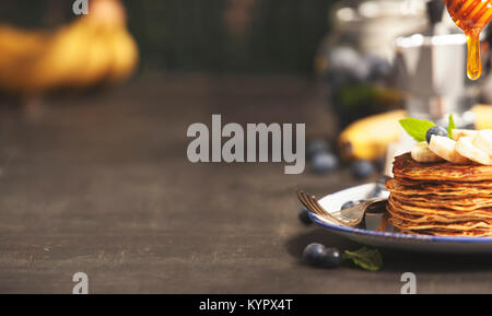 Pfannkuchen mit frischen Bananen, Blaubeeren und Honig auf einem rustikalen Tisch Stockfoto