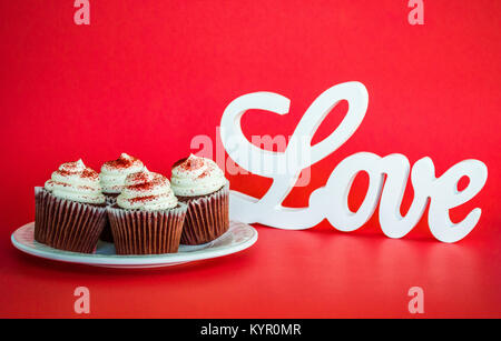 4 chocolate Cupcakes mit weißer Glasur auf einem Schild neben einem Schild, Liebe auf einem roten Hintergrund mit Kopie Raum für den Valentinstag Stockfoto
