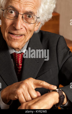 Ungeduldig alter Mann auf seine Armbanduhr zeigt. Mit Blick auf die Kamera mit einem Blick der Verzweiflung auf seinem Gesicht. Trägt einen Anzug. Nahaufnahme Stockfoto