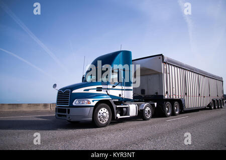Zeitgenössische Big Rig blue Semi Truck mit hoher Tag cab und überdachte Auflieger für Lieferungen Massengüter auf der breiten Autobahn mit blauer Himmel Stockfoto