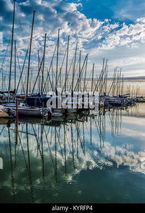 Ruhige See, Hafen und Pier bei Sonnenaufgang. Hafen mit Segelbooten und Pier in der Dämmerung mit dramatischen Wolken im Wasser spiegelt, Plattensee, Ungarn. Stockfoto