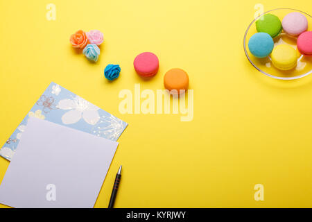 Flatlay von Notebook, bunten Kuchen Makronen, Blumen auf gelb Tabelle. Grußkarte. Platz für Text Stockfoto