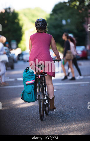 Elegante Frau in Helm und roten Kleid und Stiefel aus Leder Fahrten Fahrrad mit grün Beutel geformt zu Road Bike befestigt entlang Urban city street Stockfoto