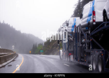 Ein leistungsstarkes Auto hauler Semi Truck Transport Autos auf einem speziellen Anhänger auf zwei Ebenen entlang der gewundenen Landstraße in den Bergen bei regnerischem Wetter Stockfoto