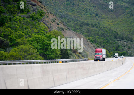 Red Big Rig Semi Truck mit Anhänger bergauf auf kurvenreichen in Mountain pass Highway mit einem anderen Lkw und Pkw Verkehr und grünen Wald Stockfoto