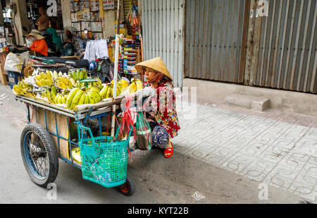 Lokale Frau mit einem konischen Hut verkauft Bananen am Straßenrand von einem Schubkarren: Blick auf die Straße in Vorstadt Saigon (Ho Chi Minh City), Süd-Vietnam Stockfoto