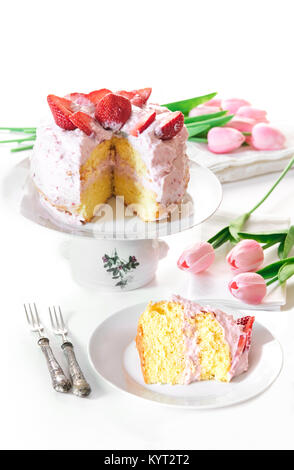 Erdbeerkuchen auf Kuchen stand mit einer Schicht bereit, auf die Platte, um bedient zu werden, alle auf einem weißen Tisch mit rosa Tulpen eingerichtet. Stockfoto