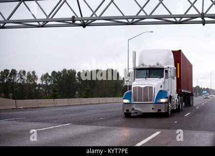 Mächtigen amerikanischen Big Rig Semi Truck mit blauen Kotflügeln Transport red Container auf Flachbett Auflieger auf regnet Multiline highway Stockfoto